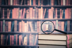 A stack of books holding a magnifying glass in front of a book shelf looking for hidden messages.