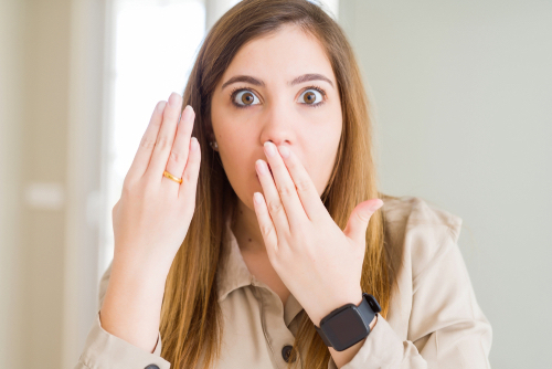 A surprised woman holding up her hand with a wedding ring on while putting her other hand to her mouth when hearing about wedding mistakes.